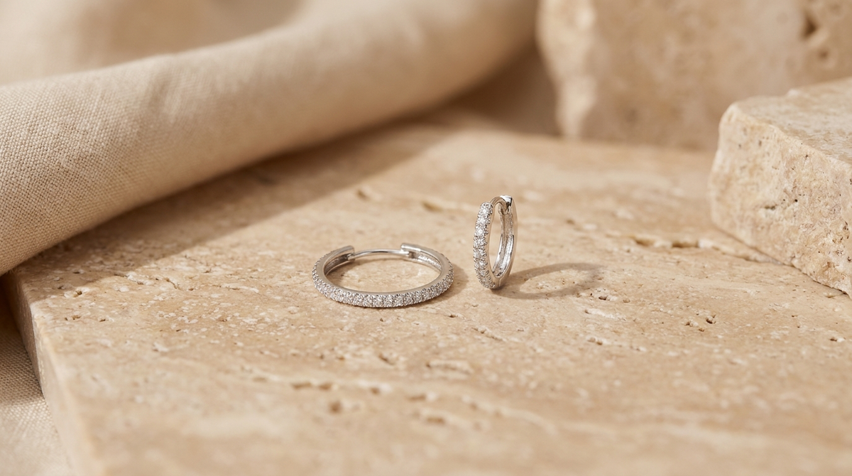 A slim diamond hoop earring beside a tiny diamond huggie earring on a warm beige surface